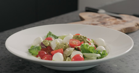 add pesto to salad with mixed greens, tomatoes and mozzarella in white bowl on concrete countertop