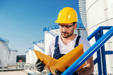 Young hardworking Caucasian unshaven refinery worker in working clothes with helmet on head and protective glasses holding folder with documents.
