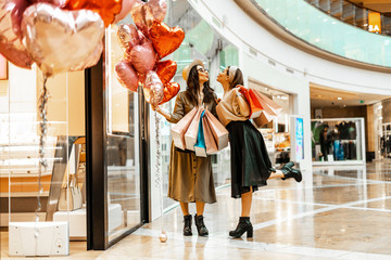 Obraz premium Shopping and entertainment, mall inside. Two beautiful girls with paper bags in the mall, heart-shaped balloons. The joy of consumption, Gift shopping, holiday.