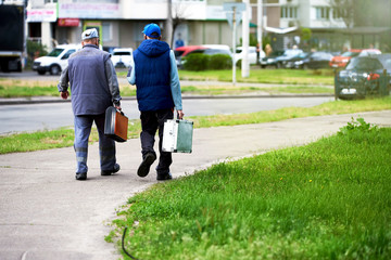 Two employees of the plumber company walk with suitcases down the street, inside the road. People go to work. Old workers, retired.