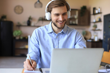 Confident businessman in earphones is writing notes or financial report while sitting at desk with...