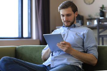 Smiling handsome man holding and using tablet with social networks at home, happy freelancer making online shopping, sitting on cozy sofa at living room