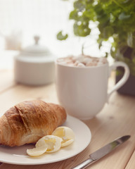 Fresh croissant with butter on a white plate and hot chocolate with melting marshmallow in a white cup on wooden table. Breakfast at home. Morning relaxation.