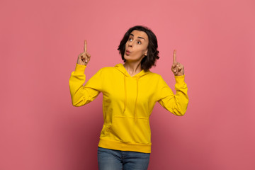 I have an idea! Close-up photo of joyful young girl in yellow sweatshirtl, who is pointing upwards with her both hands and whistling.