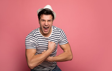 Flexing. Close-up photo of a strong man in a striped t-shirt and white hat, who is looking in the camera, yelling, and showing biceps.