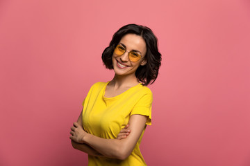 Unusual style. Close-up photo of a charming young girl in a yellow t-shirt and yellow glasses, who is looking in the camera with folded arms and smiling.