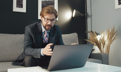 Businessman sitting on sofa in front of laptop gesticulating talking with someone through internet