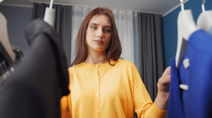 Young absorbed brunette choosing clothes at home, view from behind garment rack, low angle