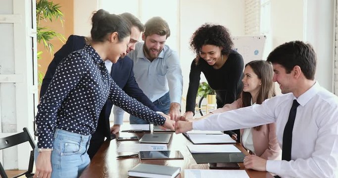 Happy multiracial business people partners gathered at office desk, bumping fists, showing team unity. Excited young mixed race colleagues celebrating success victory, demonstrating help support.