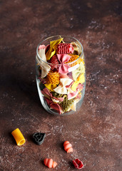 colored italian pasta of different shapes in a glass jar on a brown texture background