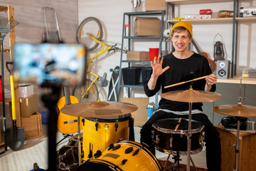 Happy young man with drumsticks sitting by drum set in front of smartphone camera
