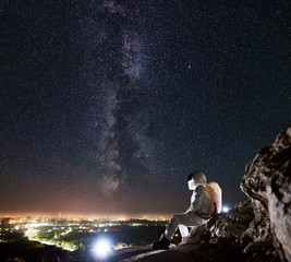 Side view of space traveler sitting on rocky mountain and enjoying the view of beautiful starry sky, Milky way, night city in valley. Concept of cosmonaut among the stars, space travelling