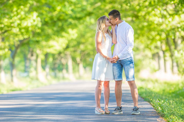 Obraz premium Young couple in love holding hands and kissing on a sunny summer day standing on a road surrounded by green trees