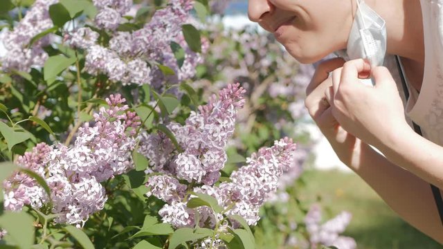 Young Caucasian woman removes the protective mask on your face and smelling the lilac flowers. Rejoices and enjoys the smell of spring and flowers. The end of quarantine. 4K