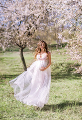 Obraz na płótnie Canvas beautiful pregnant woman in a long light dress walks in a blooming spring garden.