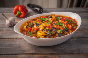 Homemade vegetable stew of eggplant, zucchini, tomatoes, peppers, garlic, prunes and greens on bowl on wooden background. Close up. Copy space for text