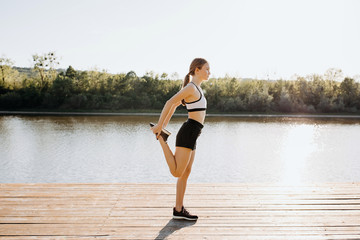 Young sportive slim fit woman stretching on a wooden platform by the lake in the morning, getting ready to run.