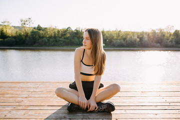 Fototapeta na wymiar Young slim fit woman resting after workout, sitting on a wooden platform by the lake, outdoors, in the morning.