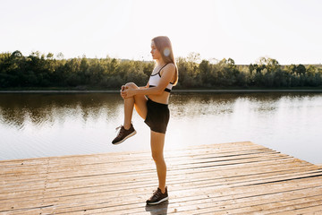 Fototapeta na wymiar Young slim fit woman doing stretching exercise in the morning. Sportive girl balancing outdoors, on a wooden platform by the lake.