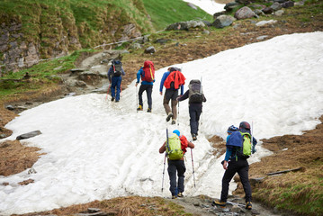 Back view of hikers with backpacks and trekking poles walking on snow covered road in mountains. Group of tourists climbing the hill while traveling together. Concept of hiking and mountaineering.
