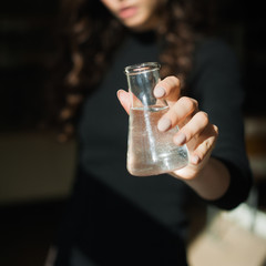 Close up portrait of young girl doctor, lab scientist, looking at the tests in the glass test tube on a white background. Focus on hands with flask with transparent liquid.