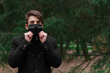 Portrait of young man in black coat wearing black medical mask. Man in classic coat walking in the park. Man in protection mask copy space. Man fixing his face mask. Person wondering in the woods.