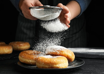 Woman decorating delicious donuts with powdered sugar at black table, closeup