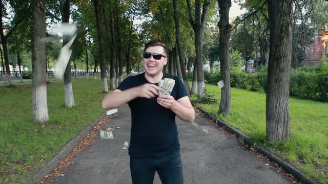 Joyful young man in glasses goes scattering money in different directions.