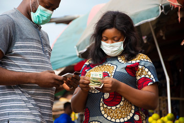 african woman in a local market wearing a face mask selling to a customer counting her money