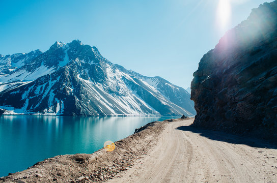 A dramatic panorama of El Yeso Dam with turquoise colour and sun flares reflecting in the water in the Andes Mountains with snow on the peaks and rock parts of the mountain exposed and a dirt road. 