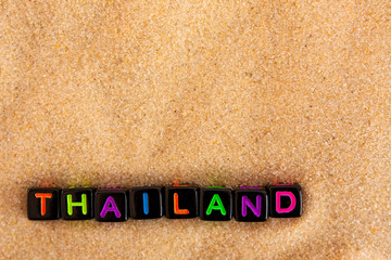 The word Thailand is made up of colored letters on black cubes on sand with a blurry sea in the background