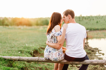 Young loving couple hug near lake sitting on log at sunset.