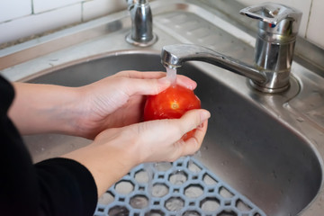 Girl is washing bright red tomato under pouring water in the sink in the kitchen. Concept of...