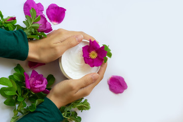Young female hands are holding jar with white anti-ageing moisturizing cream with dog rose oil essential and vitamin E   on background with bright pink dog roses, petals and green leaves