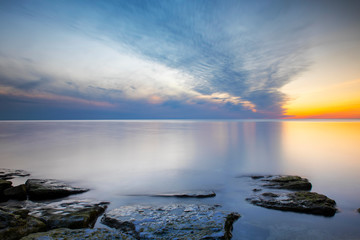 Fototapeta na wymiar Cloudy summer sunset reflecting in ocean with endless horizon and deep blue ocean, silhouette of boulders laying in the foreground in shallow water at island of Gotland, Sweden