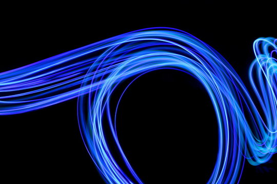 Long exposure photograph of neon blue colour in an abstract swirl, parallel lines pattern against a black background. Light painting photography.