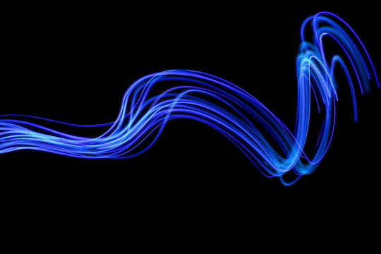 Long exposure photograph of neon blue colour in an abstract swirl, parallel lines pattern against a black background. Light painting photography.