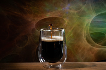 Coffee splash. Reaction of a falling drop on the hot, steaming and frothy coffee surface in a modern glass transparent cup. Studio photo with matching colored background