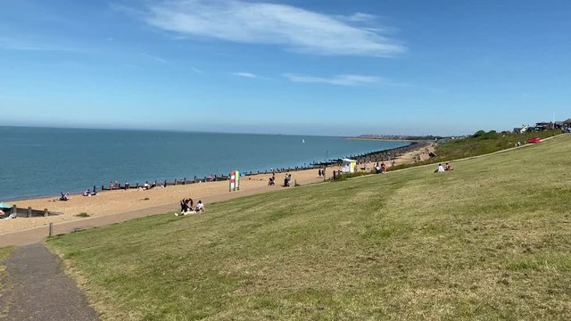 Panning shot of whitstable Beach harbour in summer on hot day with sunshine people walking on beach amd cyclist riding on pathment