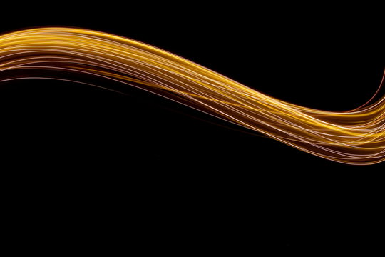 Long exposure photograph of neon gold colour in an abstract swirl, parallel lines pattern against a black background. Light painting photography.