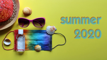 Summer vacation flat lay with protective mask painted in rainbow colors, antiseptic, beach hat, sunglasses and seashells on yellow background. Travel and coronavirus covid-19. Summer-2020 sign