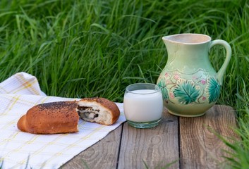 A glass of milk and a jug stand on the table against a background of green grass. Next to it, on a napkin, is a poppy seed bun.