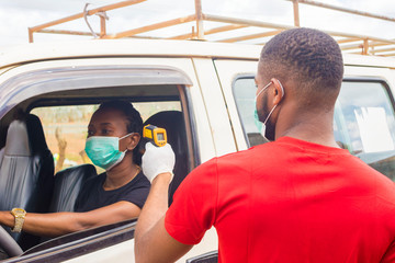 young black man wearing a nose mask, checking the temperature of a black woman driving a car