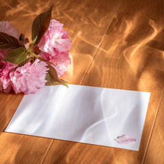 On a wooden surface lies a piece of paper and sakura flowers. Photo in pink dreamy colors. The composition is illuminated with colored light. Background Texture. Mockup, made with custom scene