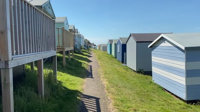 033 Panning shot of whitstable Beach harbour in summer on hot day with sunshine people walking on beach in between beach huts blue stripes and green grass