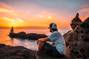 A young man sitting in a white shirt and cap looking at the orange sunset of the Pasajes San Juan...