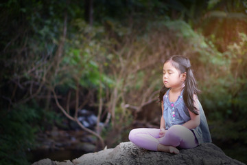 Asian child or kid girl close eye mindfulness meditating with breath and sit on stone or rock for peace and relax yoga on nature green garden and tree forest at temple park or meditate outdoor on warm