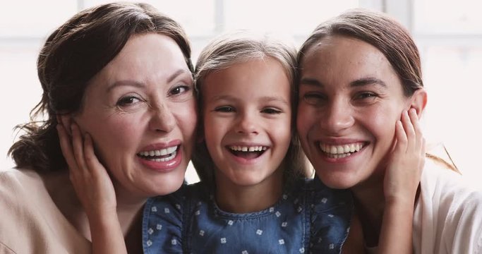 Close up portrait of happy beautiful three female generations family. Head shot little adorable preschool girl embracing smiling middle aged granny and pretty affectionate mixed race mother indoors.