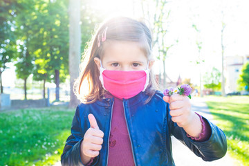 Cute little girl shows thumb up, wearing protective mask and holding bunch of wildflowers -...