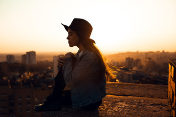 A girl in jeans and a red sweater on the roof against the background of a Sunny sunset